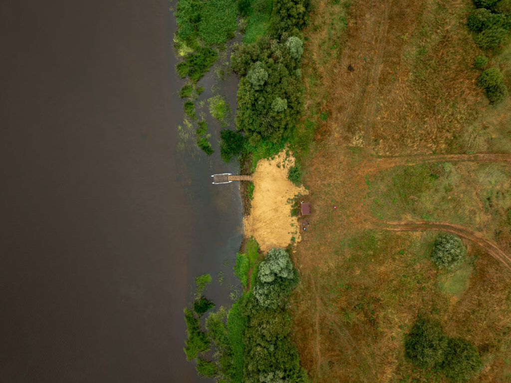 Aerial perspective of a river bordered by lush greenery, a sandy shoreline, and a small dock. A shed and dirt paths are visible in the surrounding area.