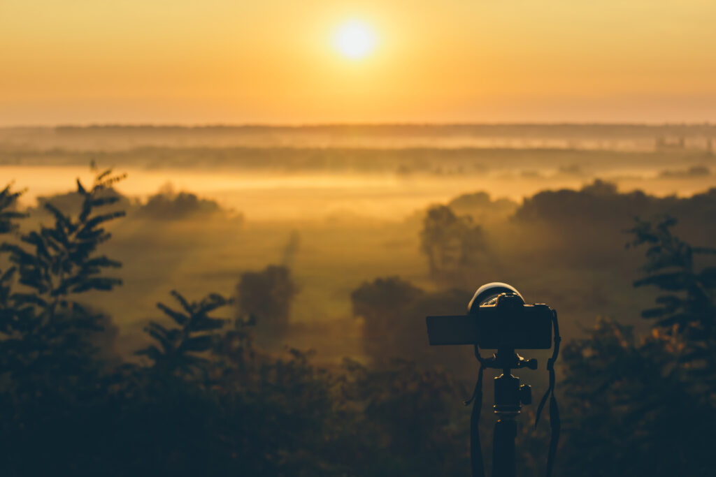 Silhouette of a camera on a tripod on a hill capturing sunrise and morning fog. Countryside landscape with camera on tripod, sunrise and morning fog.