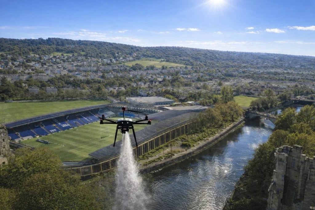 A drone sprays water over a sports field near a river and green hills under a clear blue sky.
