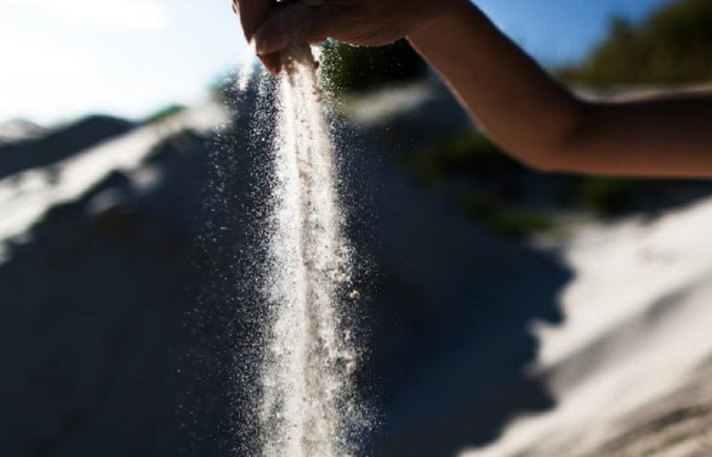 Hand scattering ashes, creating a cascade of particles against a blurred sandy background.